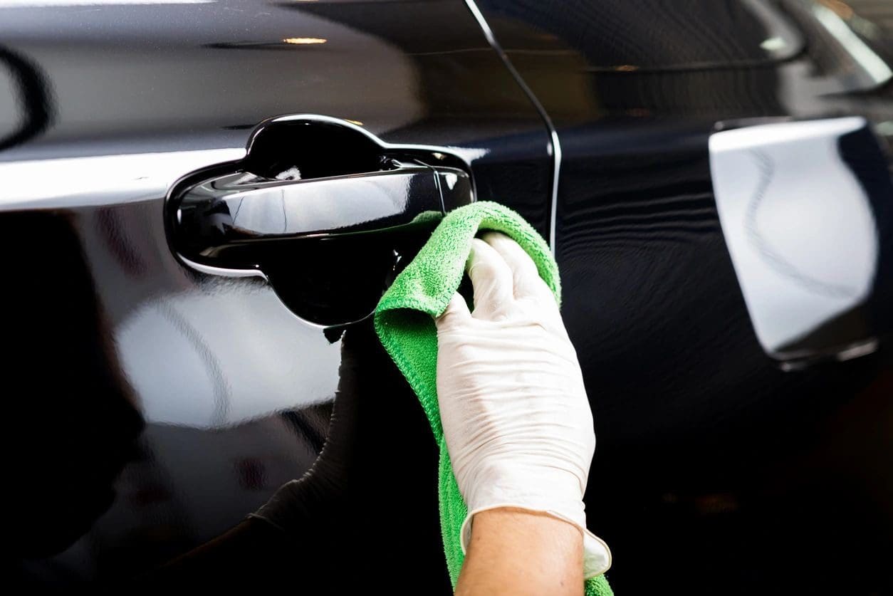 A gloved hand cleans a black car door handle with a green microfiber cloth.