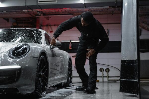 Person washing a white sports car covered in soap suds inside a dimly lit garage.